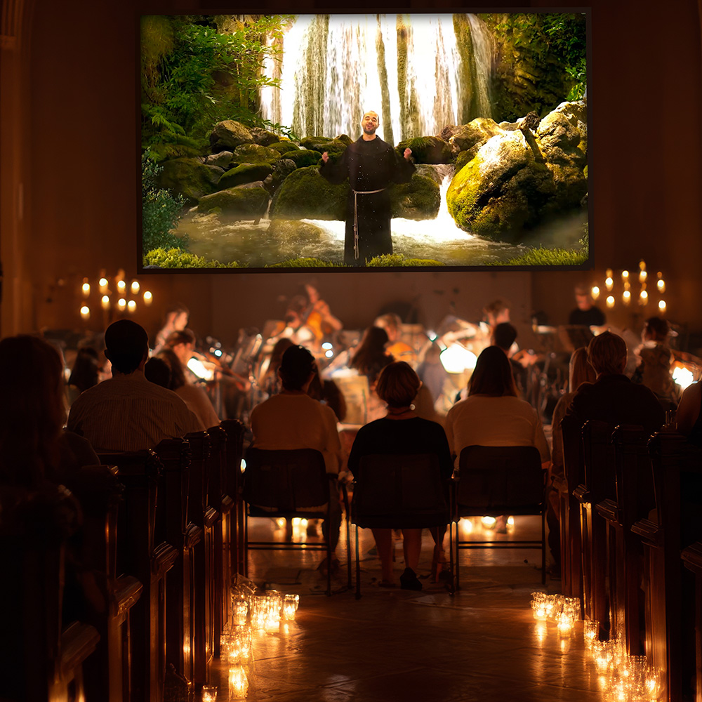 A candlelit church filled with an audience and a live orchestra, facing a large screen showing Friar Alessandro singing before a waterfall.