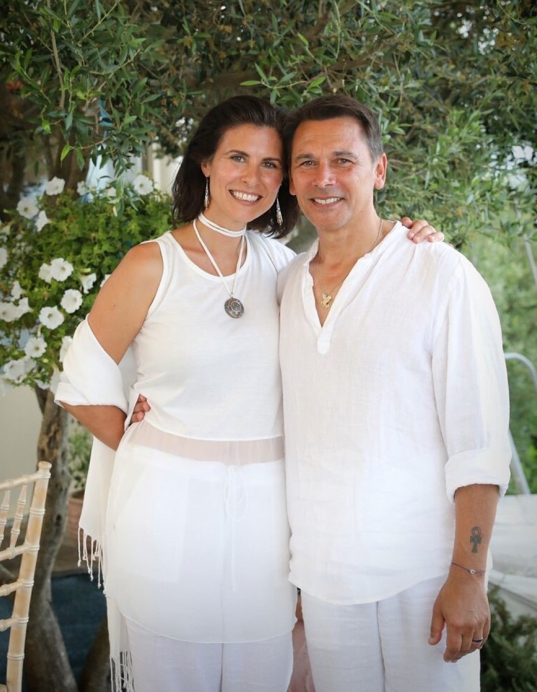 André and Gianna Ripa standing together outdoors in white linen, beneath an olive tree.